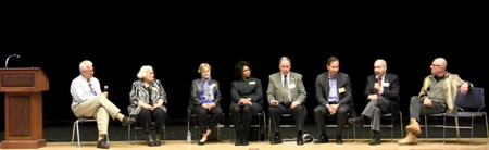 Right to left: Bobby Sager, Dr. George Holland, Daniel DiSano, Kevin Jarvis, Paula Sneed, Barbara Durgin, Marie Colantuoni Coyle, and Principal Dana Brown sit before an audience in Malden High School's Jenkins auditorium. 
